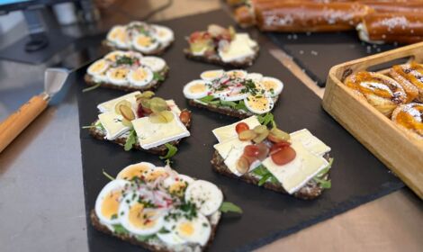 Belegte Vollkornbrote mit gekochten Eiern, Camembert, Radieschen, Trauben und frischen Kräutern, angerichtet auf einer schwarzen Schieferplatte