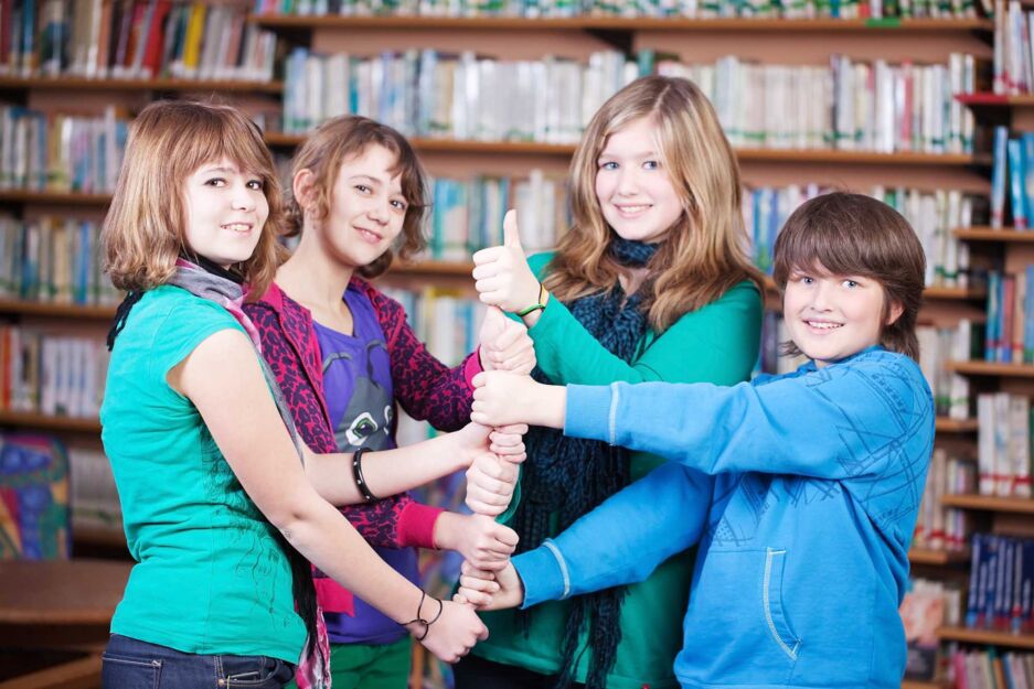 Vier Mädchen stehen in einer Bibliothek nebeneinander und stapeln ihre Handfäuste übereinander. Die oberste Hand zeigt "Daumen-Hoch".