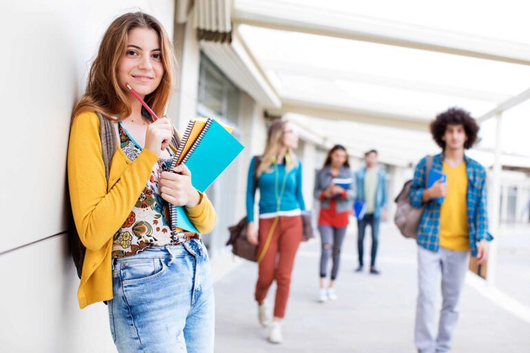 Eine Schülerin der Oberstufe steht angelehnt draußen an der Wand vom Schulgebäude und hält Collegeblöcke und einen Stift in den Armen und Händen. Im Hintergrund weitere Schüler.