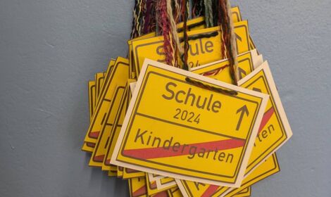 Mehrere Ortsschilder aus Papier hängen an einem Haken an der Wand. Auf dem Ortsschild in gelb steht oben Schule 2024 mit einem Pfeil nach oben und darunter durchgestrichen "Kindergarten"