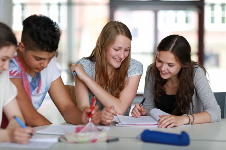 Schüler sitzen an einem Tisch nebeneinander und arbeiten in Gruppenarbeit. Sie blicken in die Hefte und halten Stifte in der Hand.