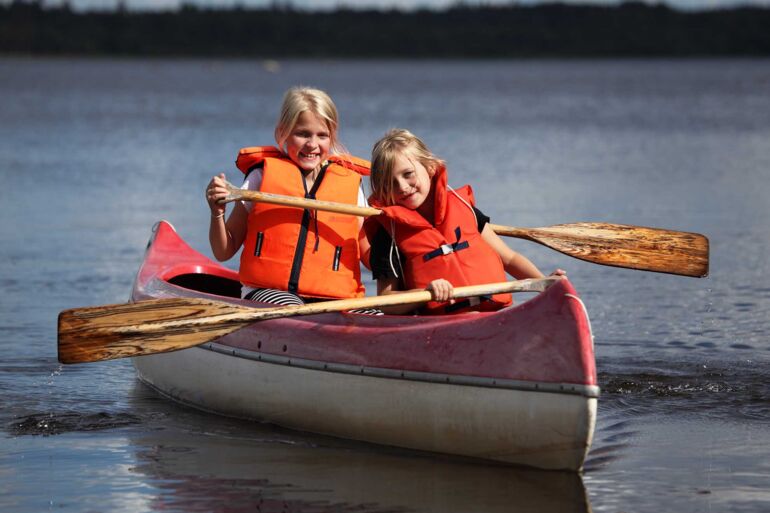 Zwei junge Mädchen mit Schwimmweste paddeln in einem Kanu auf einem See.