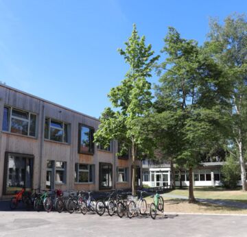 Links im Bild: Das Schulgebäude von vorne. Davor stehen Fahrradständer. Rechts eine Grünfläche mit Bäumen.