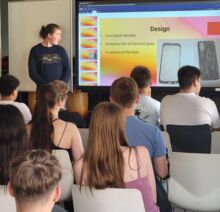 Mehrere Schüler sitzen in Reihen in einem Raum und hören einem Vortrag zu. Ein junger Schüler hält eine Präsentation auf Englisch. Ein großer Bildschirm zeigt die digitale Präsentation.