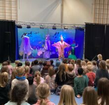 Kinder sitzen vor einer bunten Bühne mit Unterwasser-Thema, während Darsteller in fantasievollen Meereskostümen eine Theateraufführung präsentieren