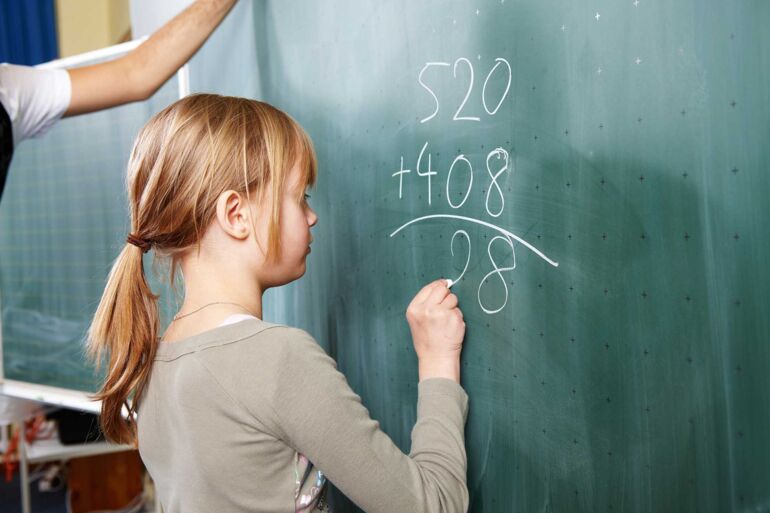 Ein junges Mädchen steht an der Tafel und errechnet eine Additionsaufgabe indem sie die Zahlen untereinander schreibt.