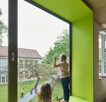 Schulkinder stehen / sitzen auf einer großen, grünen Fensterbank und blicken auf den Schulhof.