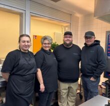 Vier Mitarbeitende eines Catering-Teams in schwarzer Arbeitskleidung stehen gemeinsam in einer Großküche vor der Essensausgabe und lächeln in die Kamera.