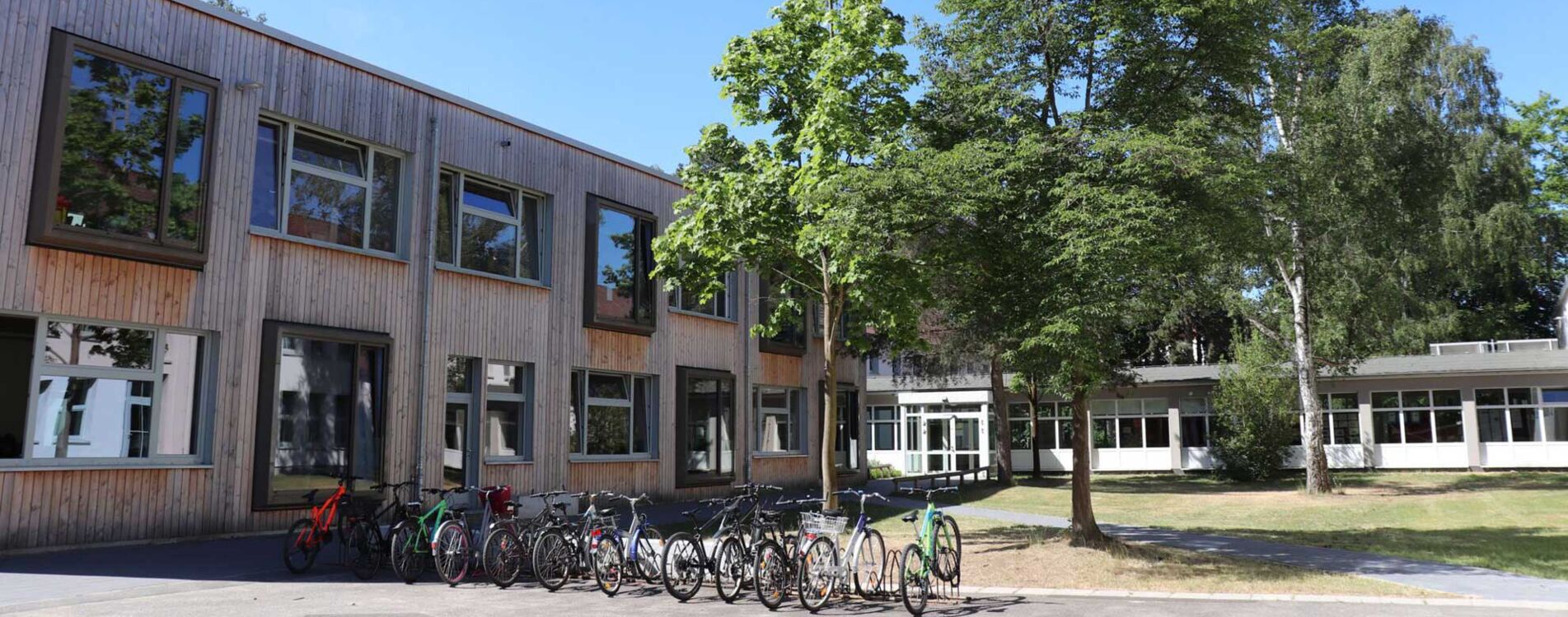 Links im Bild: Das Schulgebäude von vorne. Davor stehen Fahrradständer. Rechts eine Grünfläche mit Bäumen.