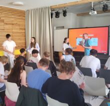 Mehrere Schüler sitzen in Reihen in einem Raum und und erhalten ihr B1 Zertifikat. Vier junge Schüler/innen halten das B1 Zertifikat in den Händen. Ein großer Bildschirm zeigt eine digitale Präsentation.