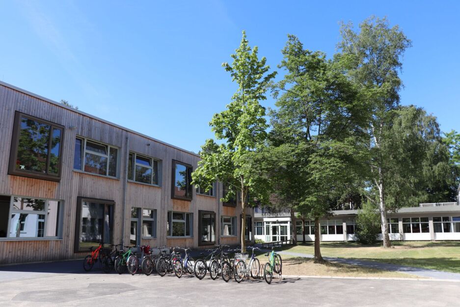 Links im Bild: Das Schulgebäude von vorne. Davor stehen Fahrradständer. Rechts eine Grünfläche mit Bäumen.