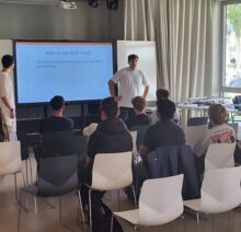 Mehrere Schüler sitzen in Reihen in einem Raum und hören einem Vortrag zu. Zwei junge Schüler halten eine Präsentation auf Englisch. Ein großer Bildschirm zeigt die digitale Präsentation.